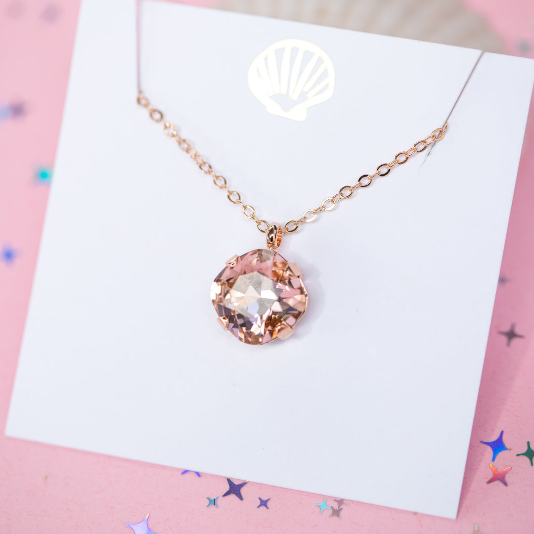 A rose gold chain necklace with a cushion cut faceted warm toned pink crystal pendant, displayed on a white card with a gold seashell logo, set against a pink background with star confetti.