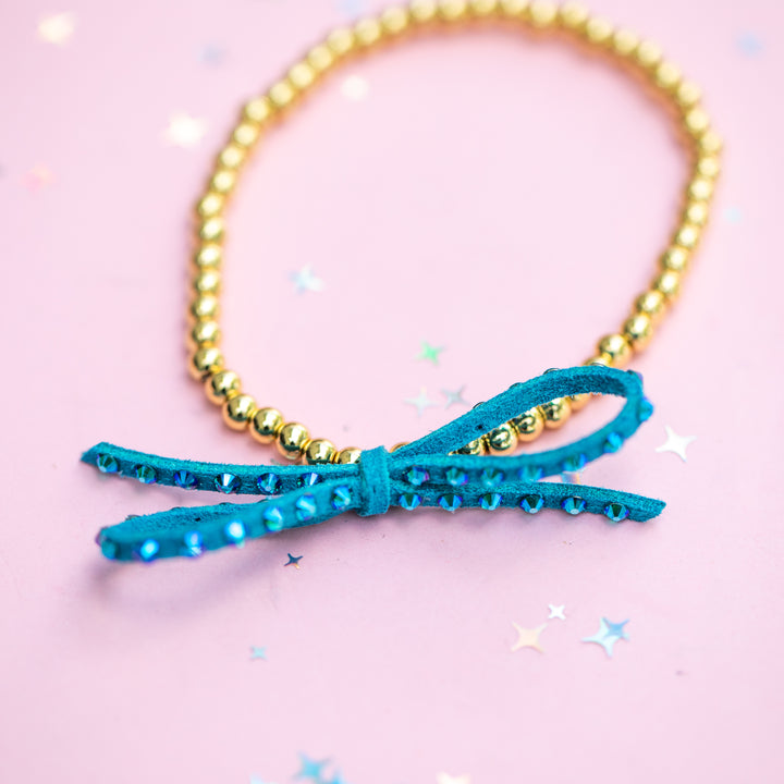A gold beaded bracelet with a turquoise blue bow charm embellished with rhinestones, set against a pink background with scattered star-shaped confetti.