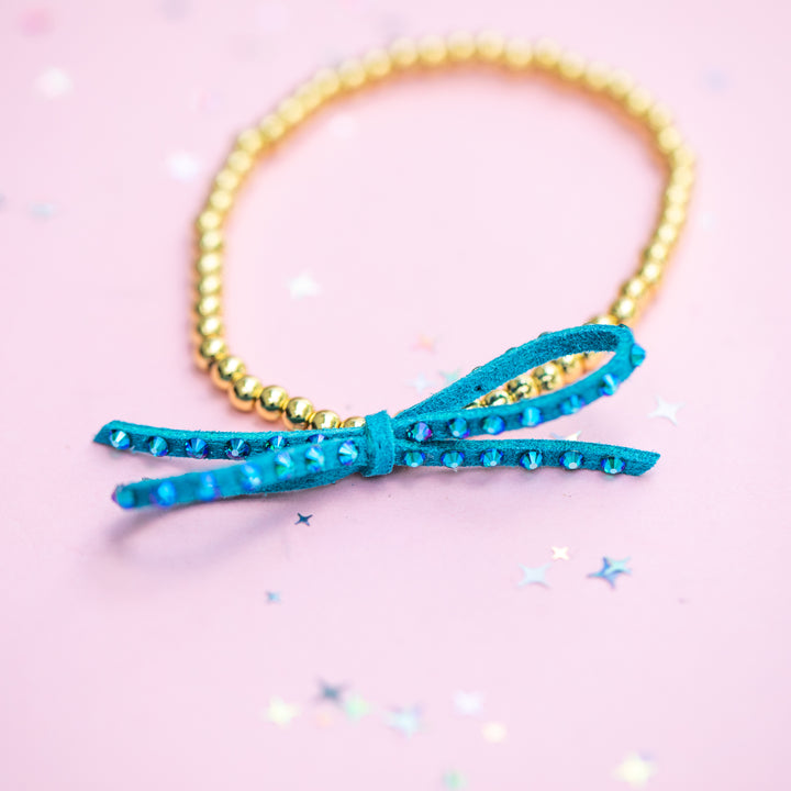 A gold beaded bracelet with a turquoise blue suede bow embellished with rhinestones, set against a light pink background with scattered star-shaped confetti.
