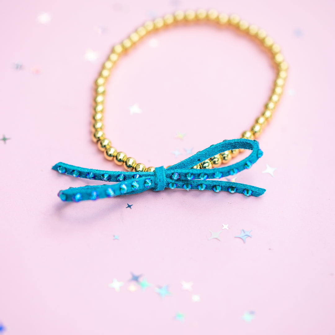 A gold beaded bracelet with a turquoise blue faux suede bow embellished with iridescent rhinestones, displayed on a light pink background with scattered star-shaped confetti.