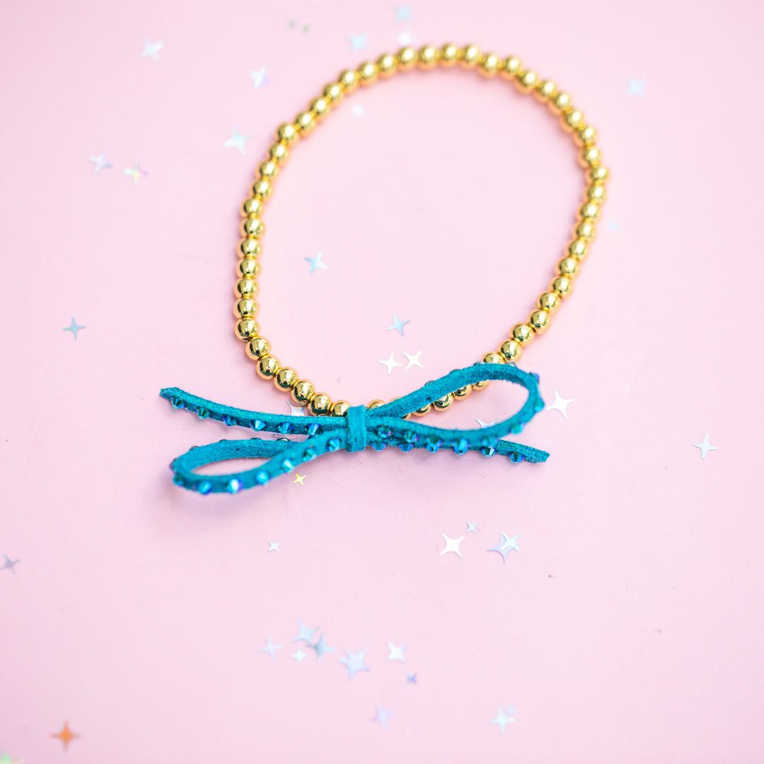 A gold beaded bracelet with a turquoise blue sequined bow charm, displayed on a light pink background with scattered star-shaped confetti.