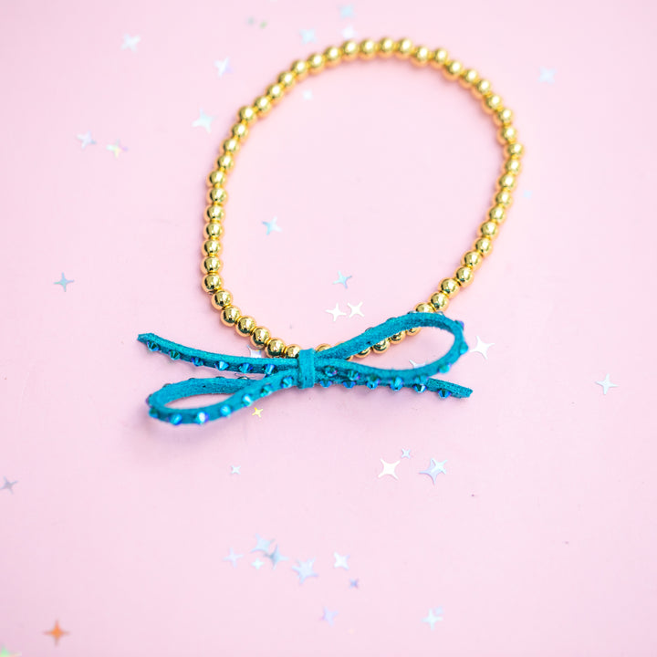 A gold beaded bracelet with a turquoise blue sequined bow charm, displayed on a light pink background with scattered star-shaped confetti.