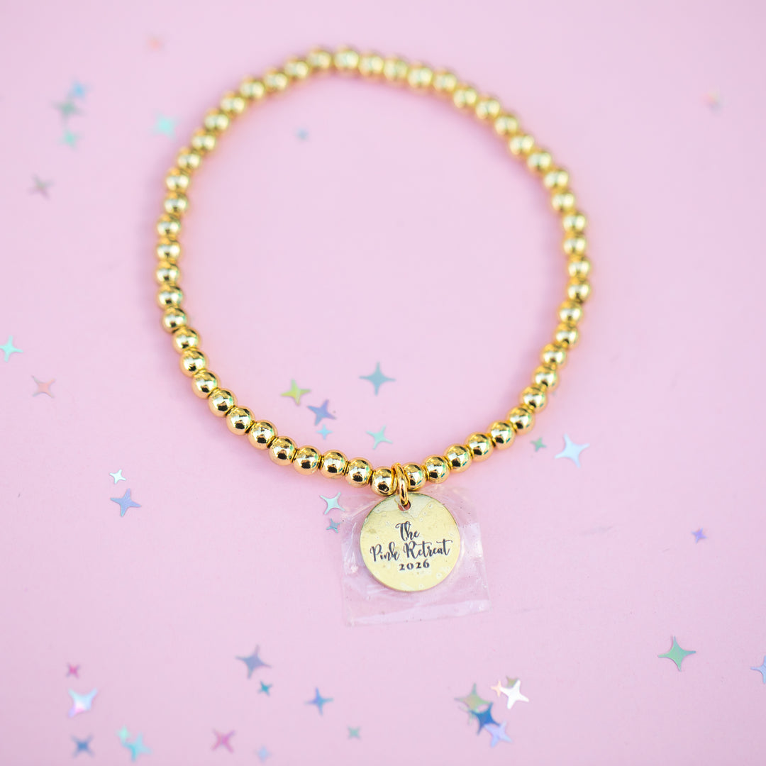 Gold beaded bracelet with a round charm on a pink background