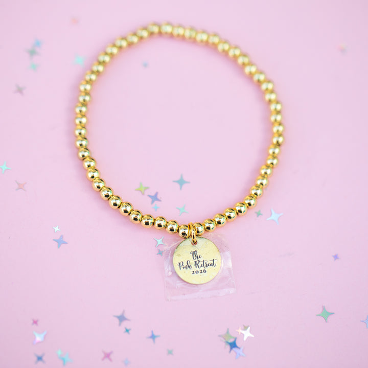 Gold beaded bracelet with a round charm on a pink background