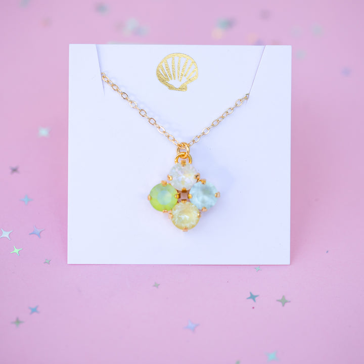 Gold necklace with green and blue gemstones on a white card against a pink background