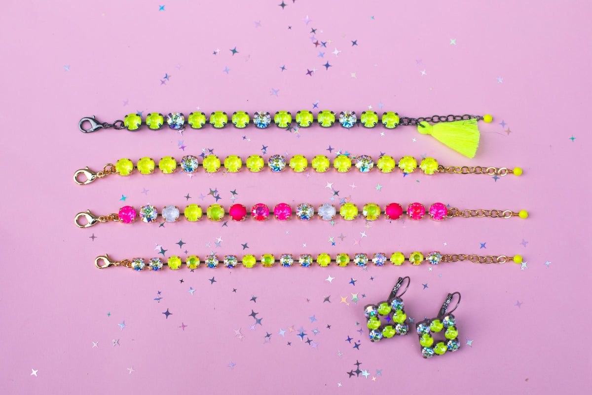 Three beaded chain bracelets and a matching pair of dangle earrings laid out on a pink background with scattered star confetti. The jewelry features neon yellow-green beads, light blue faceted beads, and pink beads, with one bracelet finished in neon yellow-green thread with a tassel.