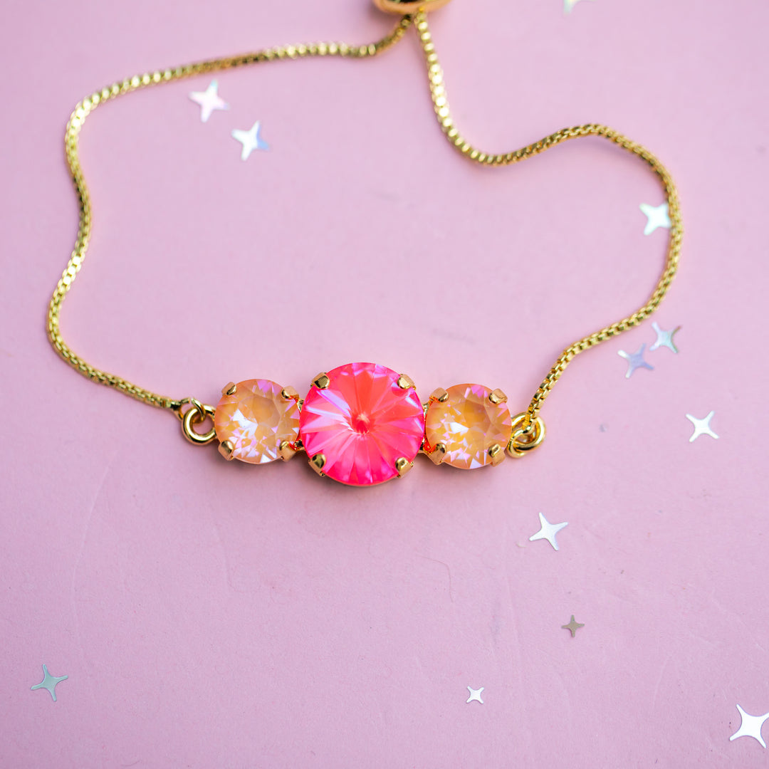 A gold chain necklace featuring a central bright pink faceted gemstone flanked by two smaller iridescent peach-orange gemstones, displayed on a light pink background with scattered silver star confetti.