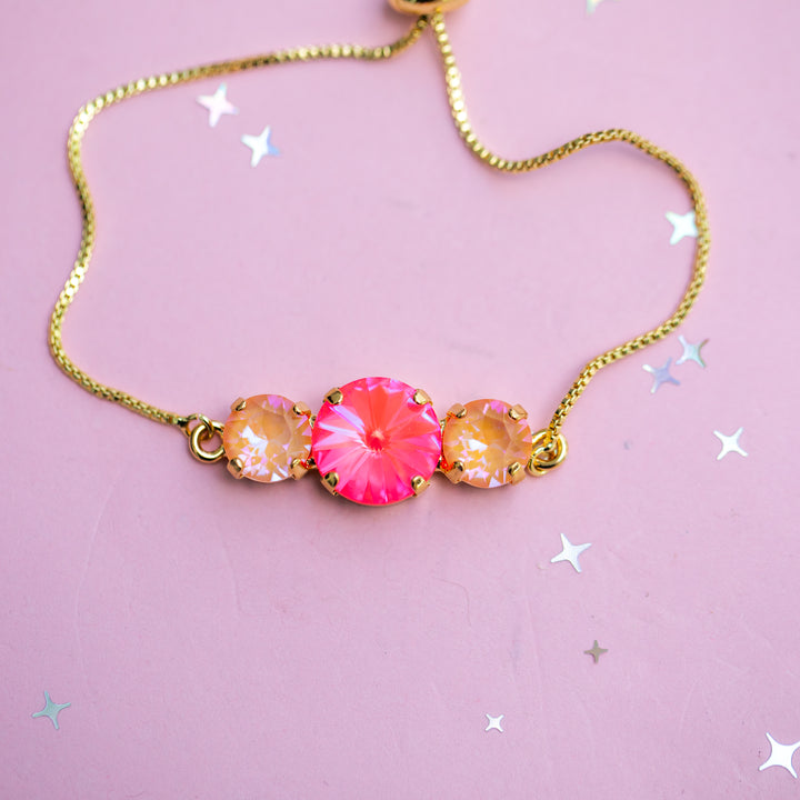 A gold chain necklace featuring a central bright pink faceted gemstone flanked by two smaller iridescent peach-orange gemstones, displayed on a light pink background with scattered silver star confetti.