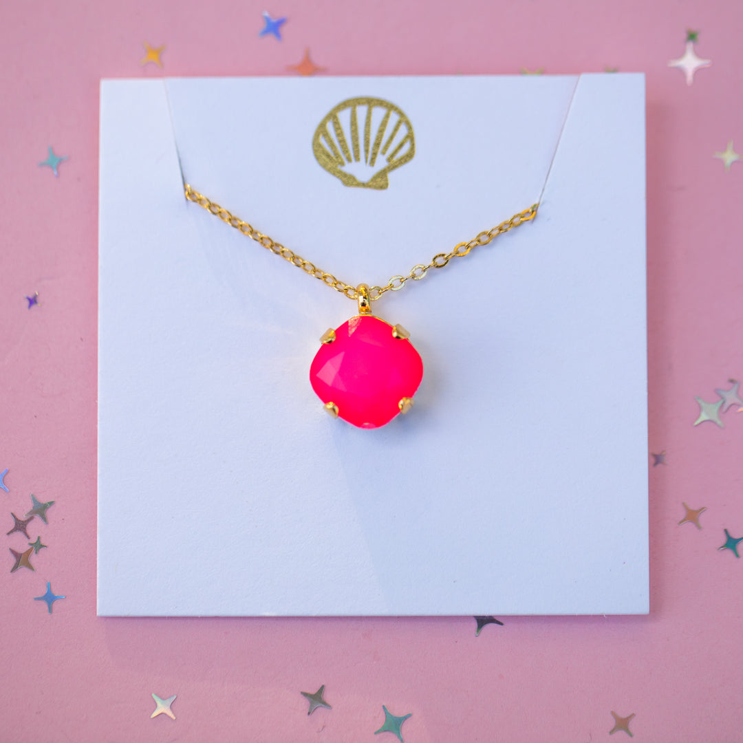 A gold chain necklace with a bright hot pink cabochon pendant, displayed on a white card with a gold shell logo against a pink background with star confetti.