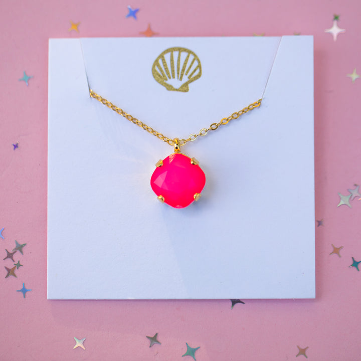 A gold chain necklace with a bright hot pink cabochon pendant, displayed on a white card with a gold shell logo against a pink background with star confetti.