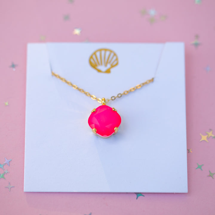 A gold chain necklace with a bright hot pink cabochon pendant, presented on a white display card with a gold seashell logo, set against a pink background with scattered star confetti.