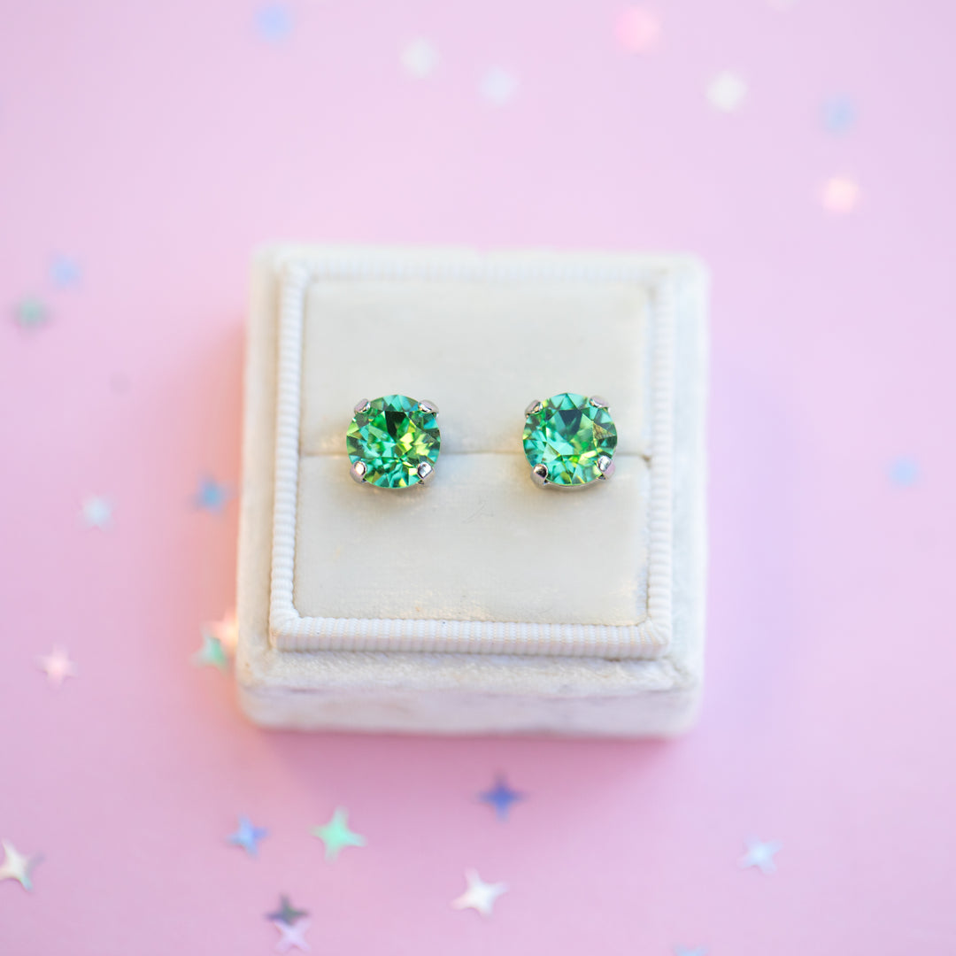 Green gemstone stud earrings in a white jewelry box on a pink background with star-shaped confetti.