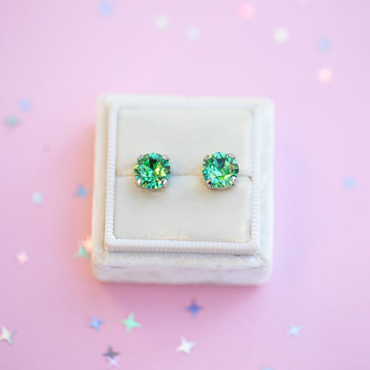 Green gemstone stud earrings in a white jewelry box on a pink background with star-shaped confetti.