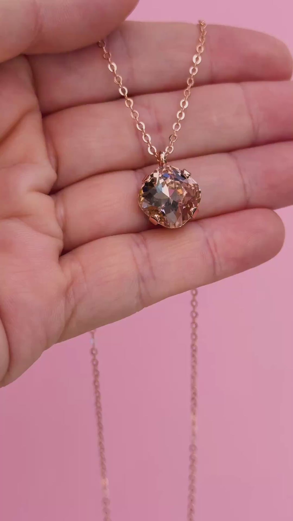 A rose gold chain necklace with a cushion cut faceted warm toned pink crystal pendant in a video shown on a hand holding it.