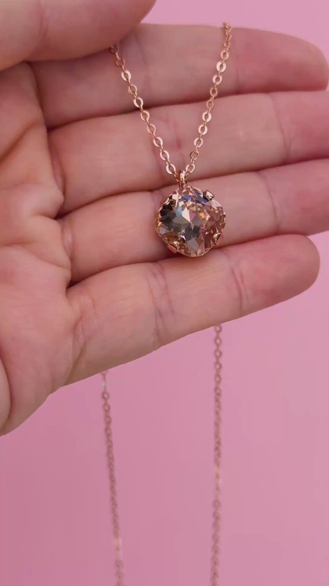 A rose gold chain necklace with a cushion cut faceted warm toned pink crystal pendant in a video shown on a hand holding it.