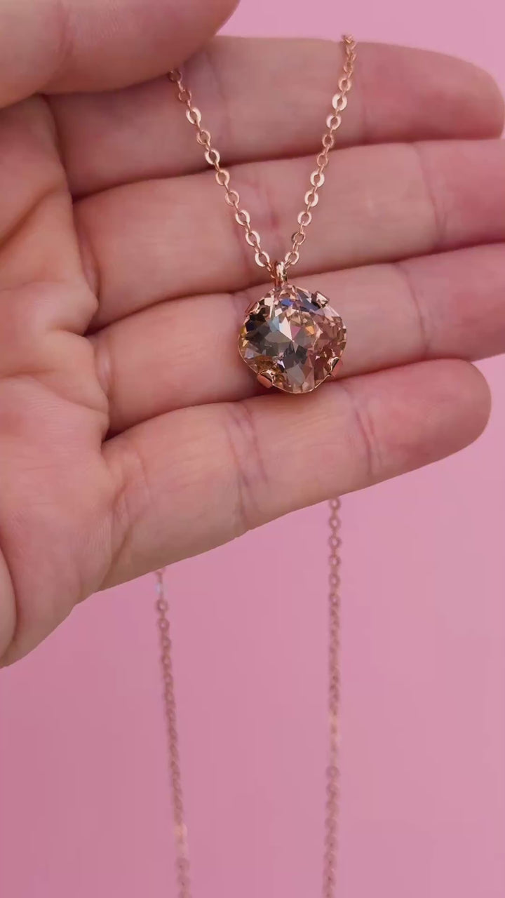 A rose gold chain necklace with a cushion cut faceted warm toned pink crystal pendant in a video shown on a hand holding it.