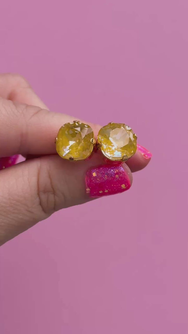 A pair of mustard yellow faceted cushion cut stud earrings held in a hand to show the size
