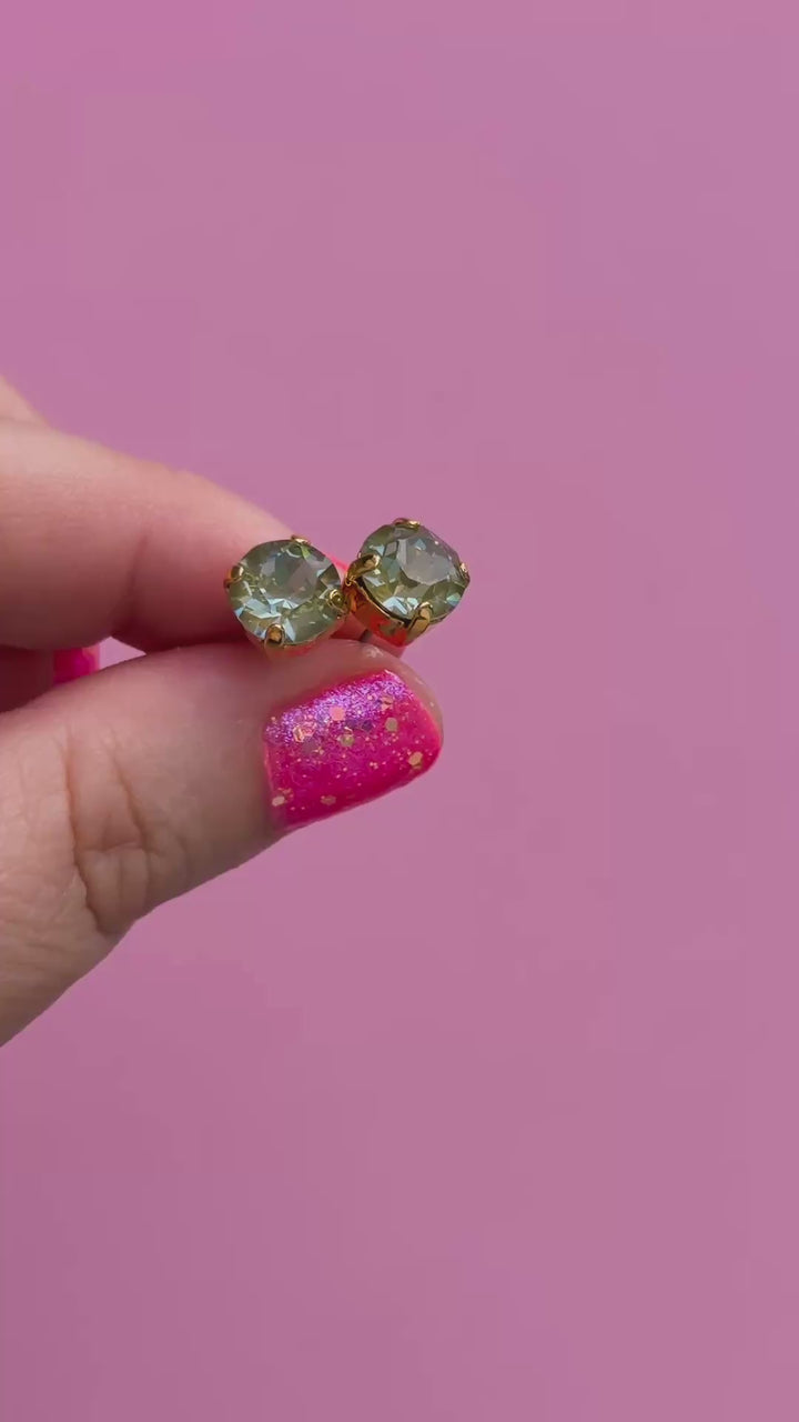 A pair of round Olive green crystal studs in a gold setting held in a hand.