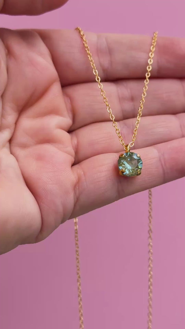A gold chain necklace with a round Olive green pendant, shown on a hand