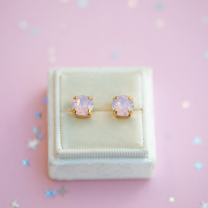 Pair of pink opal stud earrings in a white jewelry box on a pink background with star-shaped confetti.