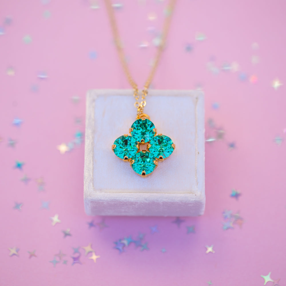 Turquoise clover-shaped pendant on a gold chain displayed on a white box with a pink background.