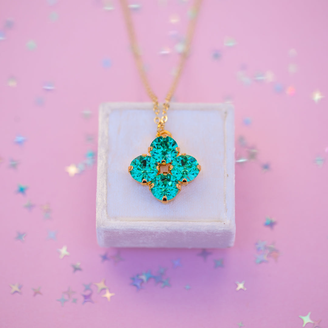 Turquoise clover-shaped pendant on a gold chain displayed on a white box with a pink background.