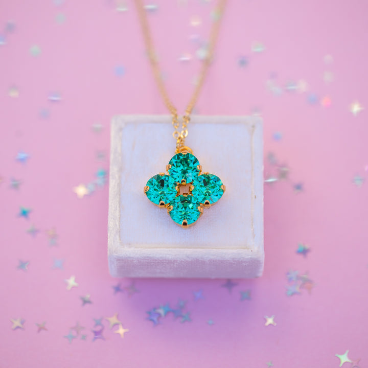 Turquoise clover-shaped pendant on a gold chain displayed on a white box with a pink background.