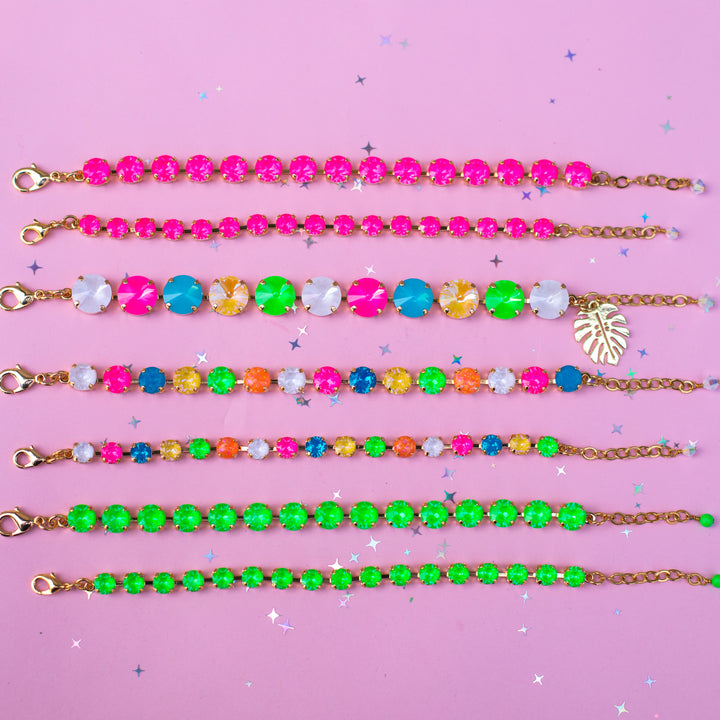 Six colorful beaded bracelets with gold-tone chains and clasps, arranged horizontally on a pink background with small star confetti. The bracelets feature various combinations of neon pink, green, blue, yellow, orange, and white rhinestones, with one bracelet featuring a gold leaf charm.