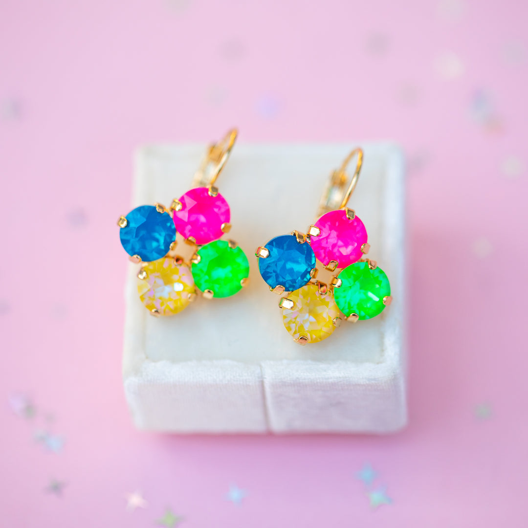 A pair of gold-tone dangle earrings with colorful gemstone clusters, displayed on a small white jewelry box against a pink background with scattered confetti.