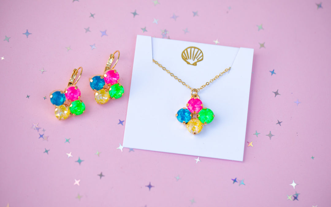 A gold necklace and matching drop earrings with colorful gemstone clusters, displayed on a white card against a pink background with scattered star confetti.