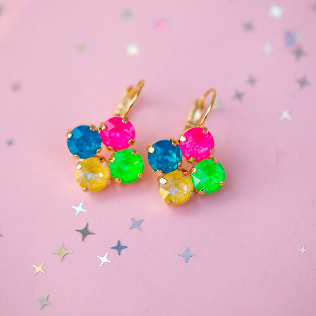 A pair of gold-tone cluster earrings with faceted gemstones in bright neon blue, pink, yellow, and green, displayed on a pink background with scattered star-shaped confetti.