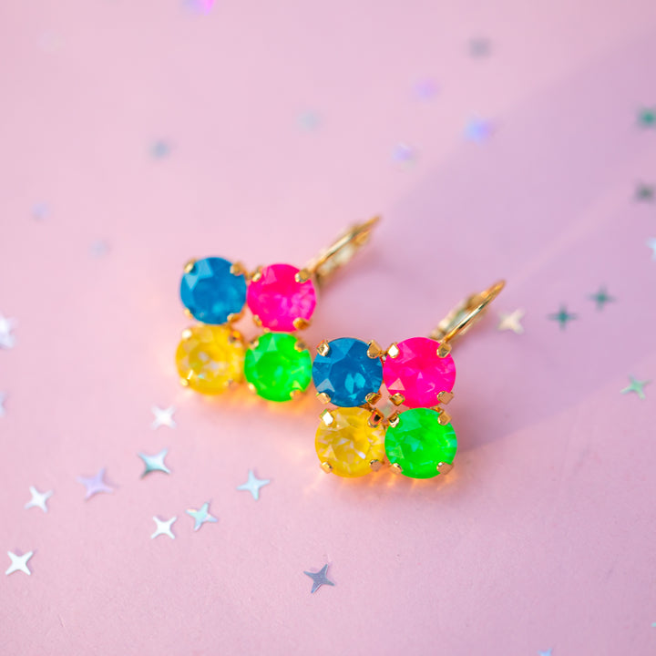 A pair of gold earrings with three colorful round gemstone clusters in blue, pink, yellow, and green, displayed on a pink background with scattered star confetti.