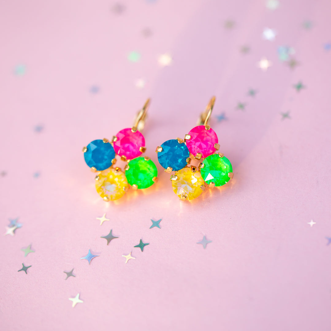 A pair of gold-tone dangle earrings with five colorful faceted gemstones in pink, blue, green, and yellow, arranged in a flower-like cluster on a light pink background with scattered star confetti.