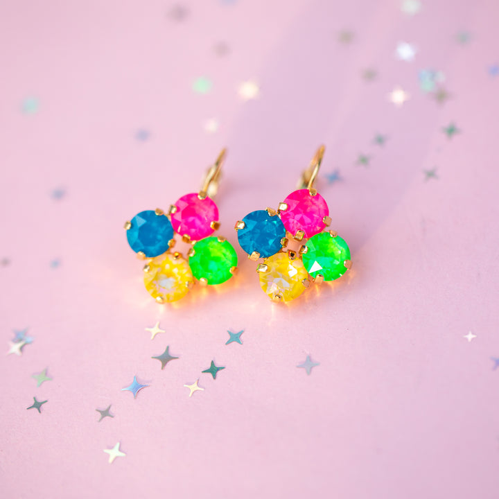 A pair of gold-tone dangle earrings with five colorful faceted gemstones in pink, blue, green, and yellow, arranged in a flower-like cluster on a light pink background with scattered star confetti.